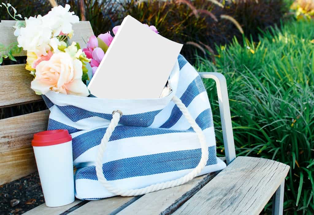 A striped beach bag filled with flowers and a book on a wooden bench outdoors.