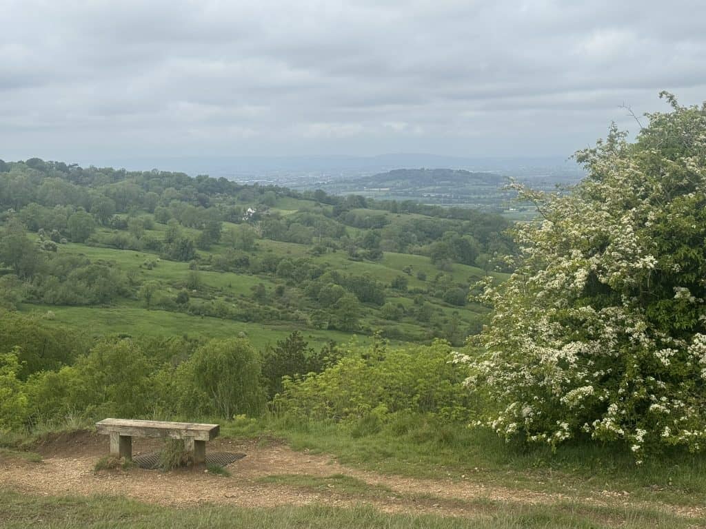 Wooden bench overlooks rolling green countryside and distant hills beneath a cloudy sky. The peaceful viewpoint offers a place to rest and enjoy the scenery along the Cotswold Way.