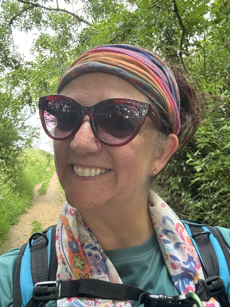 Smiling hiker wearing sunglasses, a colorful scarf, and a backpack on Day Eleven of the Cotswold Way near Bath. A shaded trail lined with green trees stretches behind her as the long-distance walk nears its end.