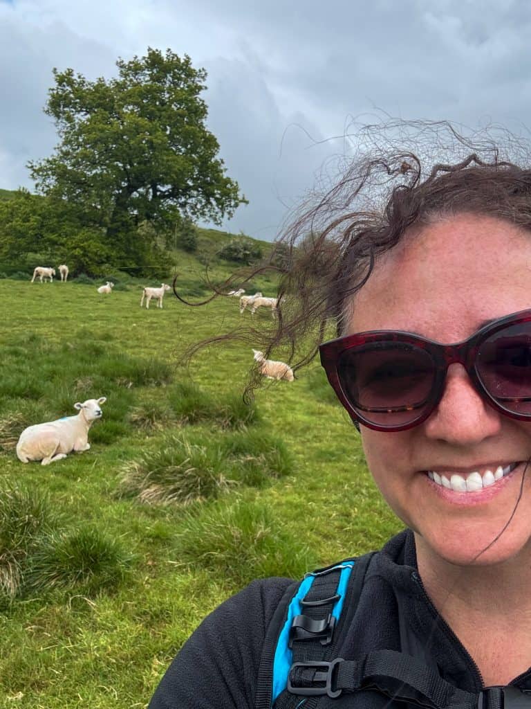 Smiling hiker wearing sunglasses and a backpack poses beside a grassy hillside dotted with sheep on Day Two of the Cotswold Way. Dark storm clouds gather overhead while sheep graze and rest in the field behind her.