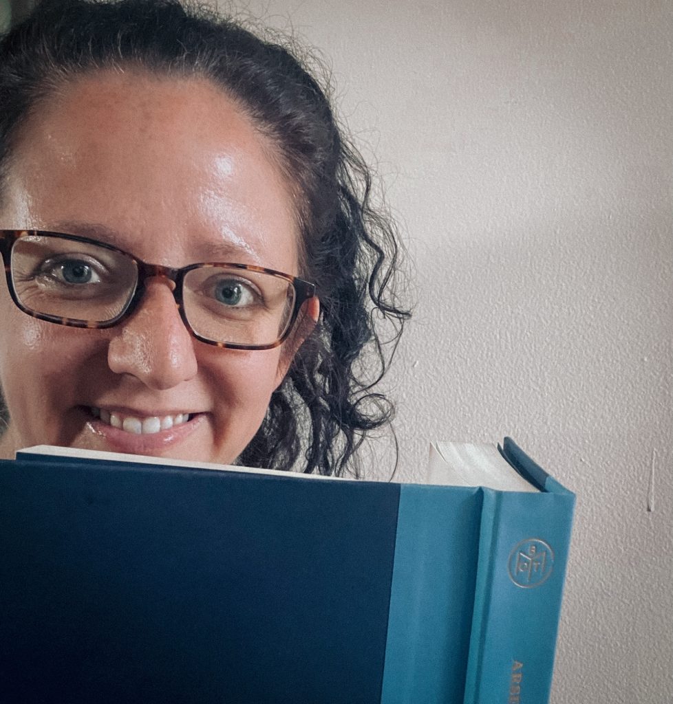 A woman with curly dark hair and glasses smiles while holding up a large blue hardcover book. The book partially covers her face, revealing only her eyes and the top of her smile. The background is a simple, neutral-colored wall, and the lighting is soft and natural.