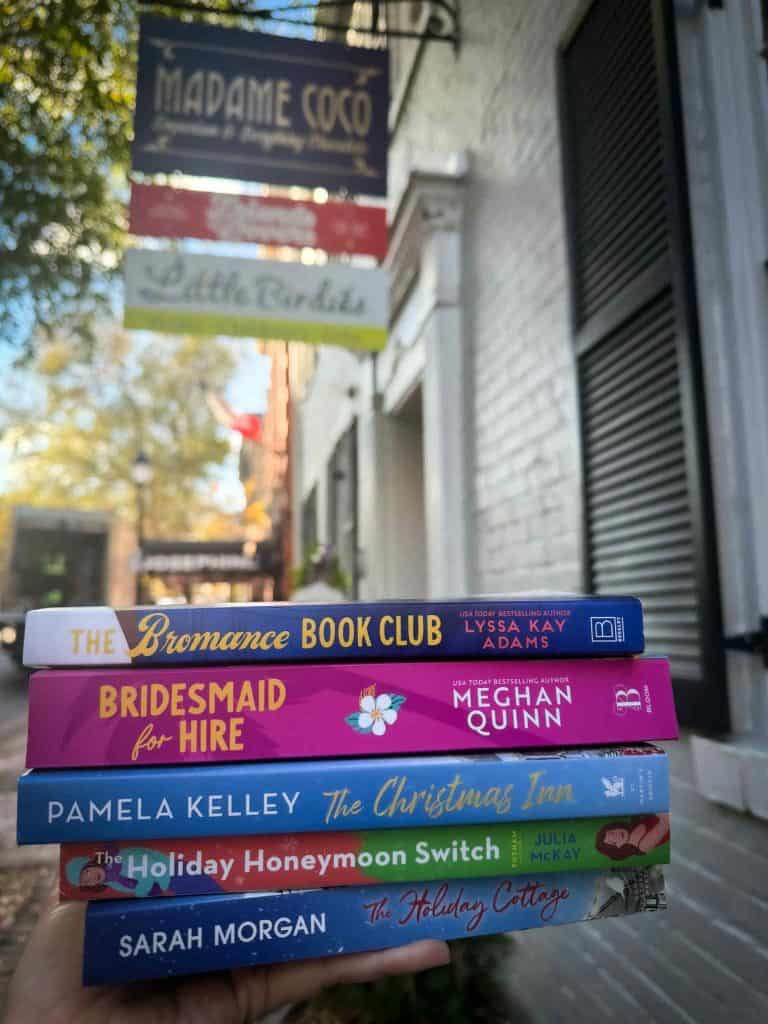 A hand holds a colorful stack of romantic novels in front of boutique signs, promoting fun and easy-to-read books that inspire readers to read more.