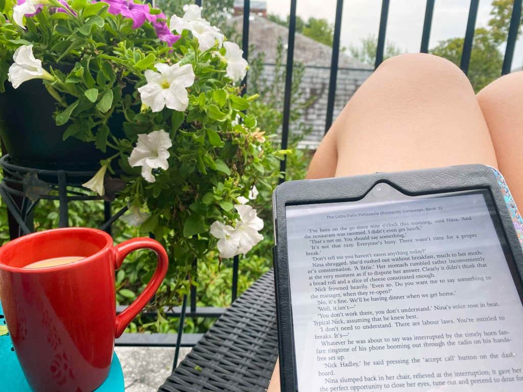 A person relaxing on a balcony with a red mug of coffee, a flowering plant nearby, and an e-reader displaying a novel, creating a peaceful atmosphere that encourages readers to read more.